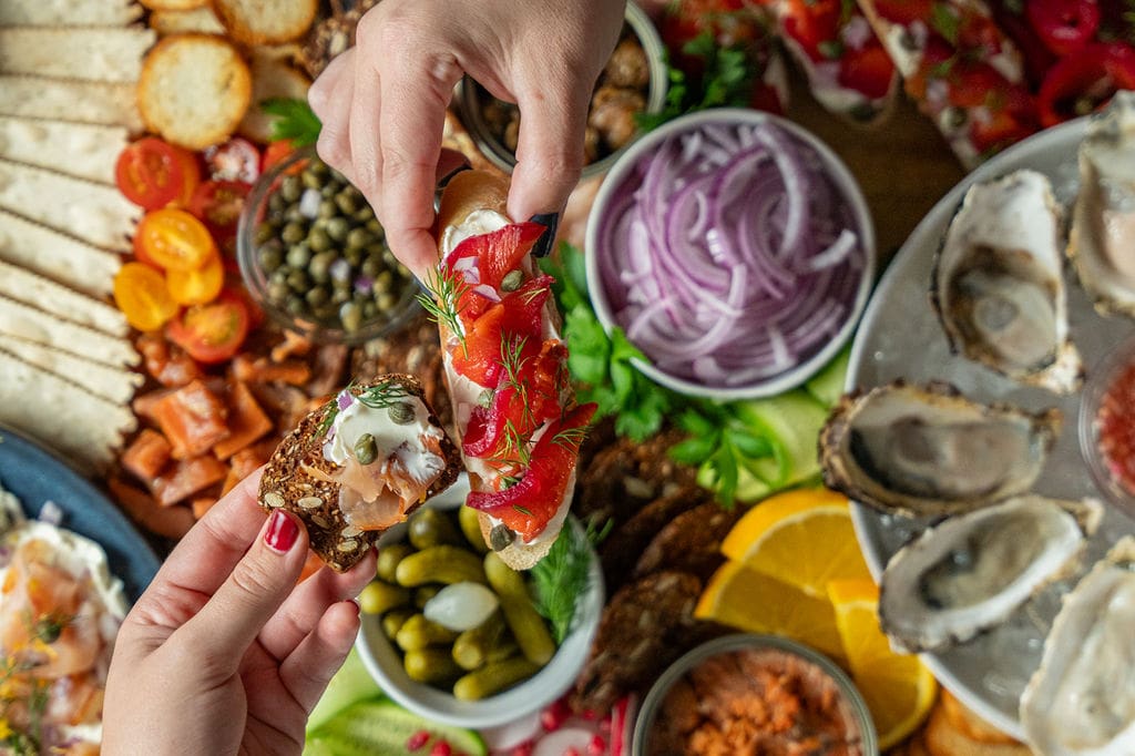 A seafood charcuterie board adorned with fresh oysters, tinned seafood, smoked salmon, lox, bread, crackers and produce. Above the charcuterie board, two hands hold a prepared bite.