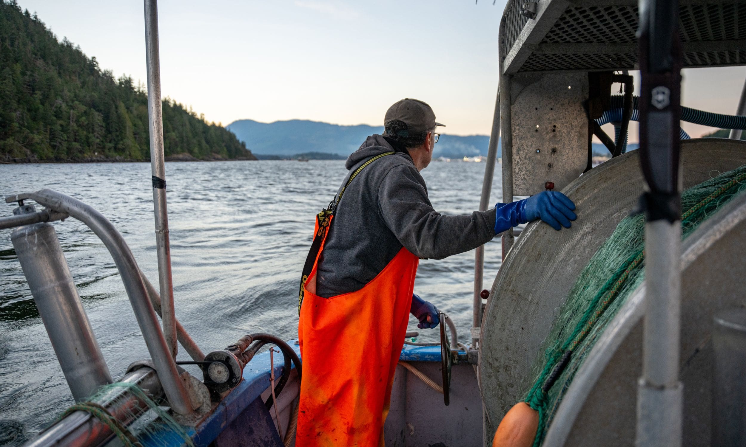 A fisherman aboard his boat, looking out over the water and mountains in the distance.