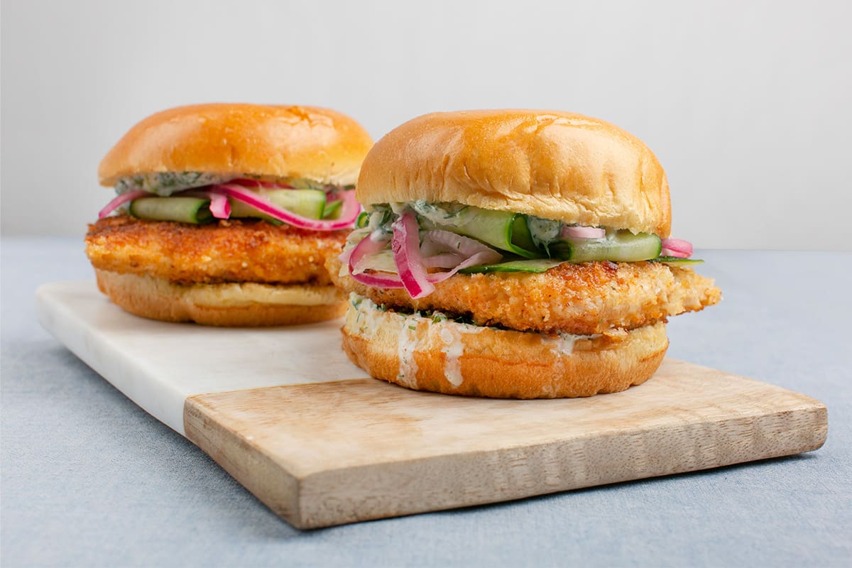 Two fish burgers on a serving board, made with breaded pike fillets, cucumber, red onion and tzatziki.