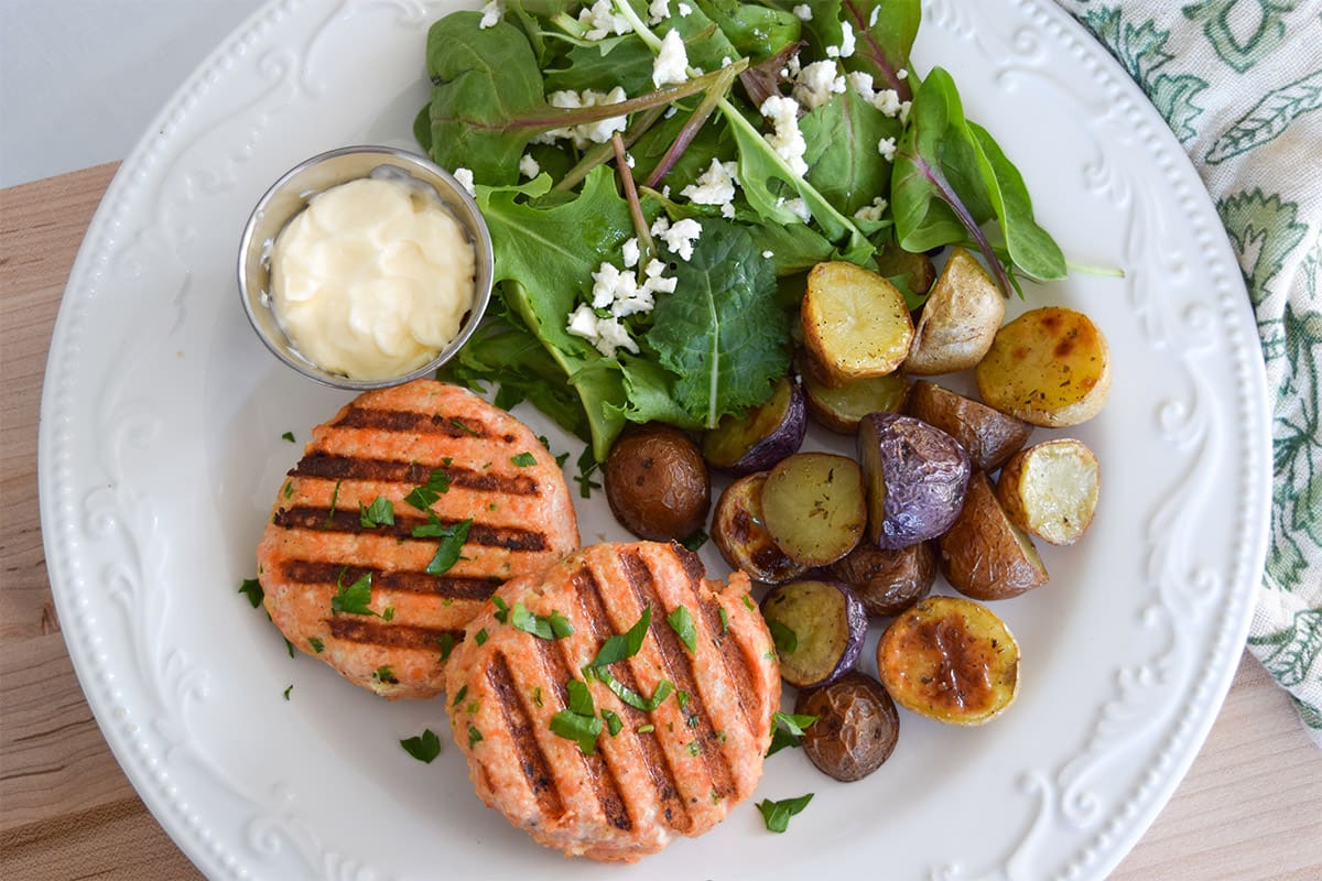 Two grilled coho salmon patties served on a white plate with roasted baby potatoes, salad greens, and aioli.