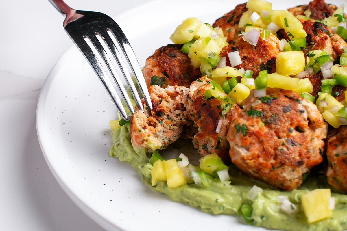 Seasoned sockeye salmon balls are served on a white plate with a green avocado sauce and pineapple salsa.