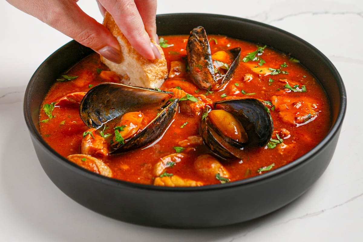 A piece of fresh baguette is being dipped into a bowl of seafood stew.