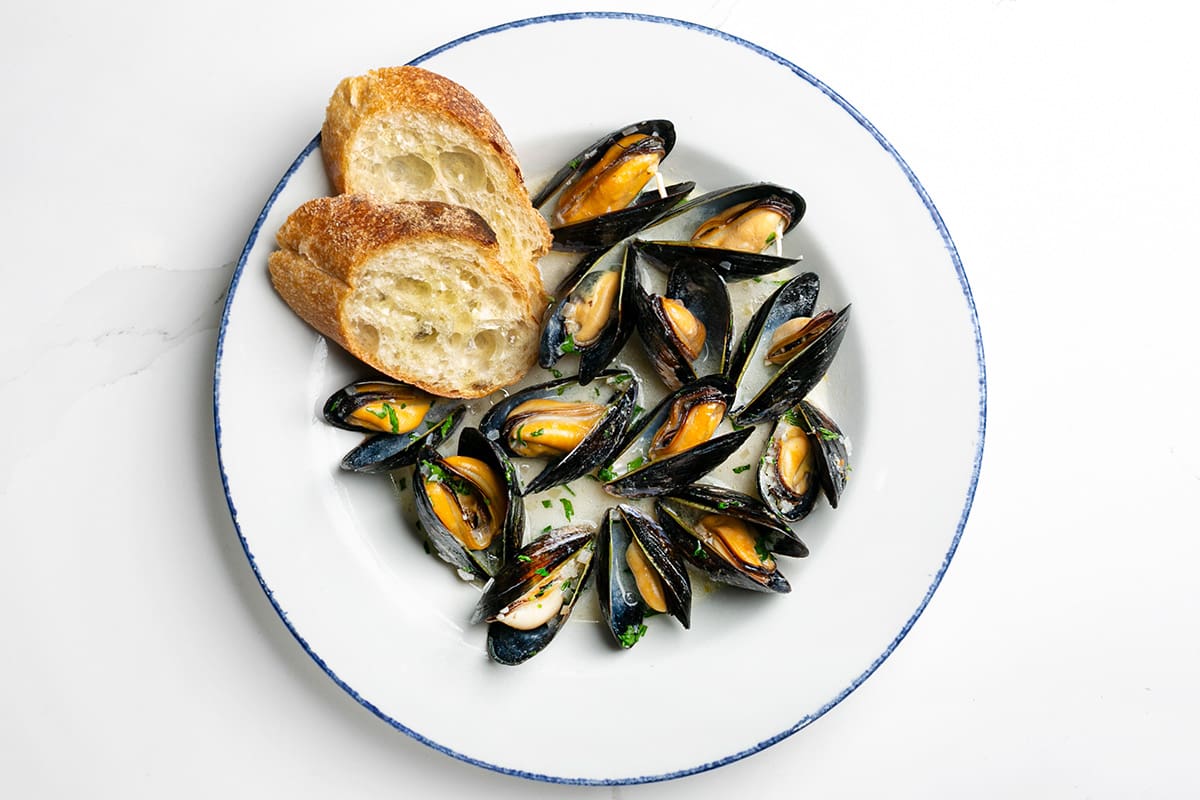 A bowl of fresh mussels cooked in a white wine and garlic broth with a side of fresh baguette.