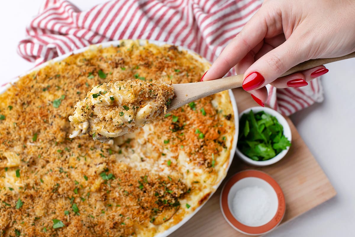 A hand scooping a serve of Dungeness crab mac and cheese from a baking dish.