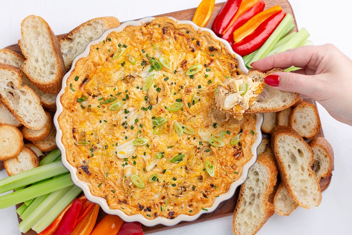 A baking dish of hot crab dip, served on a board with cut vegetables and pieces of fresh baguette.