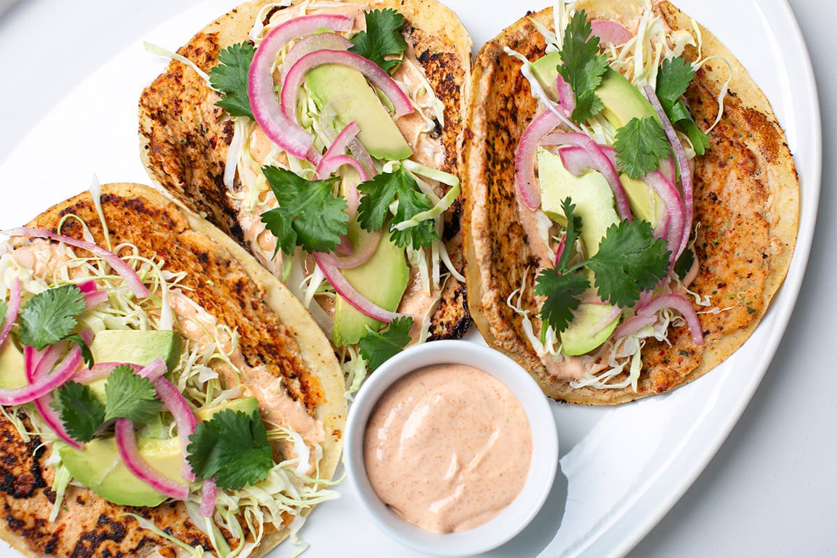 Three smashed pike tacos served on a platter with a side of sriracha sauce.